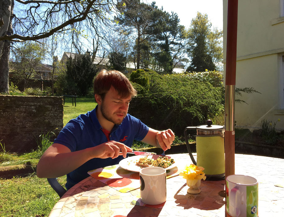 A young man enjoys a breakfast of scrambled eggs on toast and coffee sat at a table in a sunny garden during the coronavirus lockdown